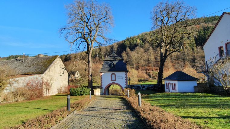 Eingangstor der Klosteranlage St. Thomas mit gepflastertem Weg, umgeben von Bäumen und Gebäuden, vor einem bewaldeten Hügel.