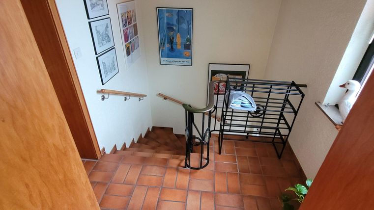 Ein heller Flur mit Fliesenboden und Holzdetails. Eine Treppe führt nach oben, und an der Wand hängen einige Bilder.