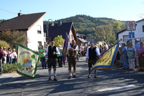Een feestelijke parade met mensen in traditionele klederdracht die vlaggen vasthouden. Op de achtergrond zijn toeschouwers en een schilderachtig landschap te zien.