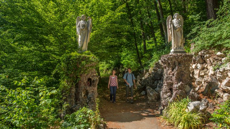 Twee mensen lopen over een bospad tussen twee standbeelden van engelen.