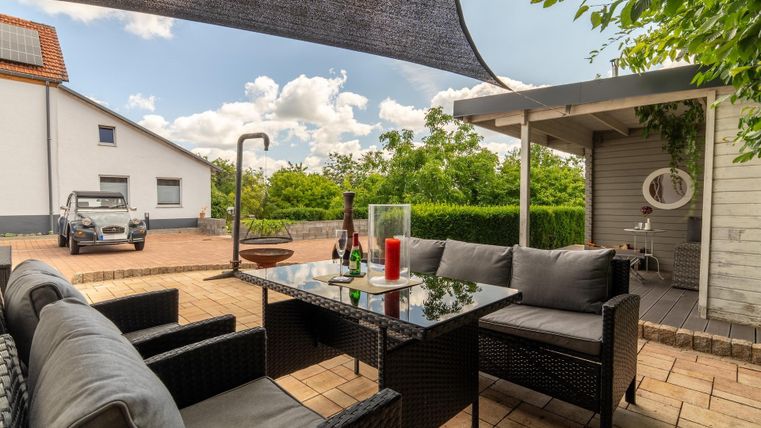 Een gezellige buitenruimte met een zwart rattan sofa en een tafel. Op de achtergrond is een verzorgde tuin en een heldere, vriendelijke sfeer te zien.