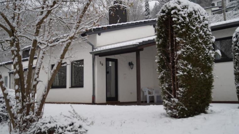 A pretty house in the snow with a snowy garden. The entrance is clearly visible, and trees frame the scene.