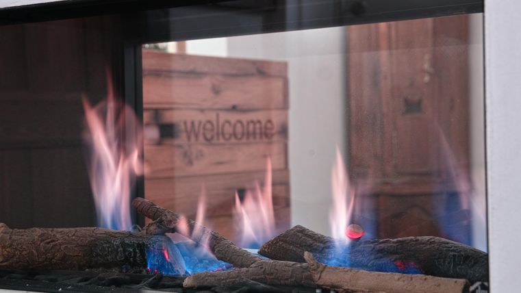 Een moderne open haard met brandende houtblokken. Op de achtergrond is een houten kist te zien met het opschrift "welcome".
