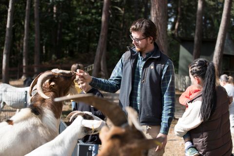 Een man voert een geit in een landelijke omgeving. Op de achtergrond staan andere dieren en mensen.