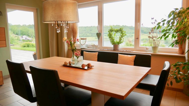 Een lichte eetkamer met een grote houten tafel en zwarte stoelen. Door de ramen kijkt men uit op een groene landschap met planten en decoraties in de ruimte.