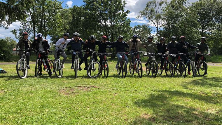 A group of ten cyclists stands on bicycles on a green meadow. In the background, there are trees and a blue sky with some clouds visible.