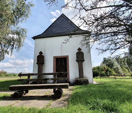 Chapelle blanche avec toit en ardoise, flanquée de deux colonnes en pierre. Un banc en bois se trouve devant, entouré d'une pelouse verte et d'arbres., © Tourist-Information Bitburger Land