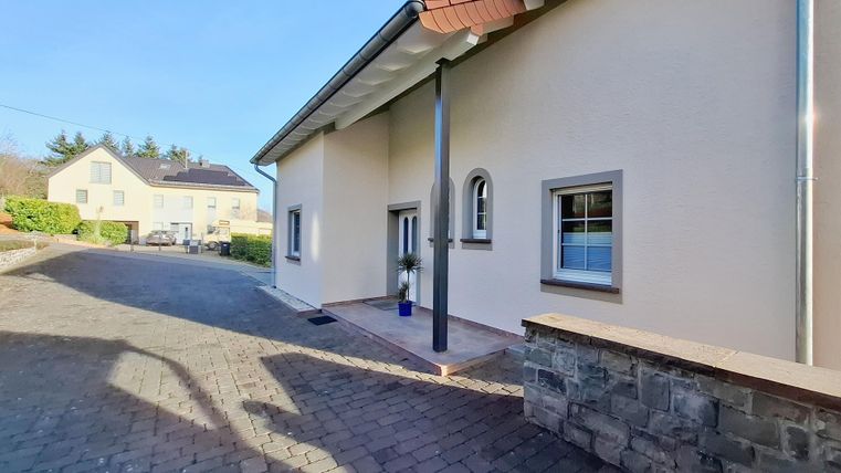 Een modern huis met een geplaveide weg en een heldere, blauwe lucht. Op de achtergrond zijn andere gebouwen zichtbaar.