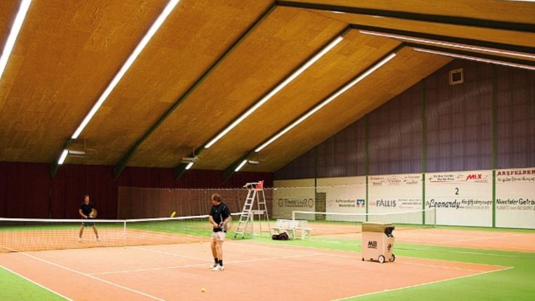 Een moderne tennisbaan met een oranje vloer en groene tapijt. Twee spelers trainen binnen.