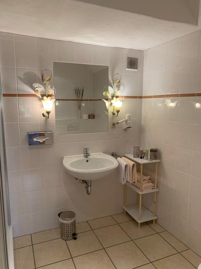 Een moderne badkamer met een wastafel, een spiegel en discrete wandlampen. Er zijn handdoeken en verzorgingsproducten op een kleine planken.