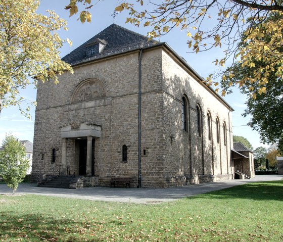L'église Saint-Hubert à Wolsfeld, construite en pierre claire, se trouve dans un environnement automnal avec des arbres et une pelouse verte., © Tourist-Information Bitburger Land