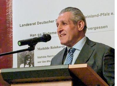 Een man staat bij de microfoon en spreekt tijdens een evenement. Op de achtergrond zijn teksten en een afbeelding te zien.