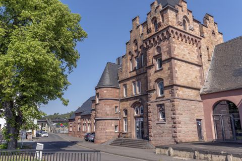 Een historisch gebouw in de neogotische stijl met torens en rode bakstenen. Op de achtergrond zijn bomen en een helder blauwe lucht te zien.