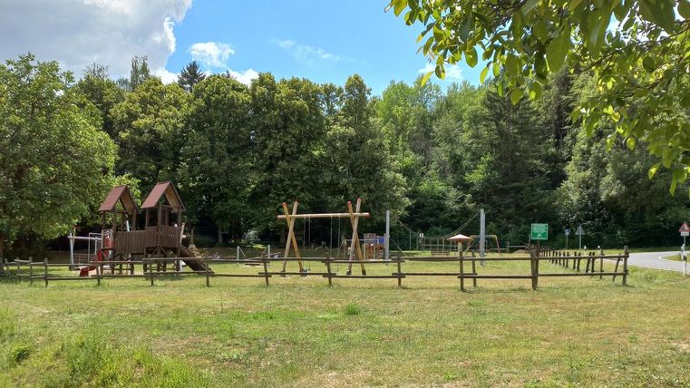 Playground with wooden houses, swings, and climbing frame, surrounded by trees and grass, under blue sky with clouds.