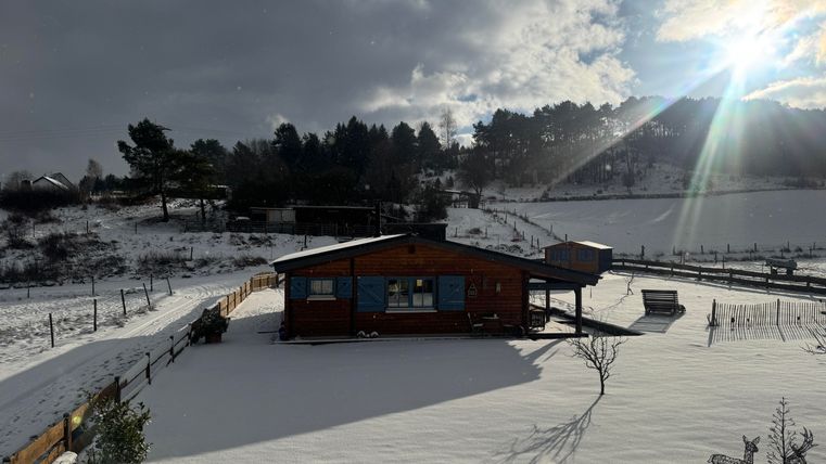 A cozy wooden house in the snow, surrounded by a winter landscape. The sun shines through the clouds, creating a picturesque atmosphere.