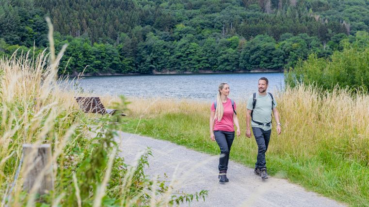 Zwei Wanderer gehen auf einem Schotterweg am Ufer des Rursees entlang. Umgeben von hohen Gräsern und bewaldeten Hügeln im Hintergrund genießen sie die Natur an einem sommerlichen Tag.