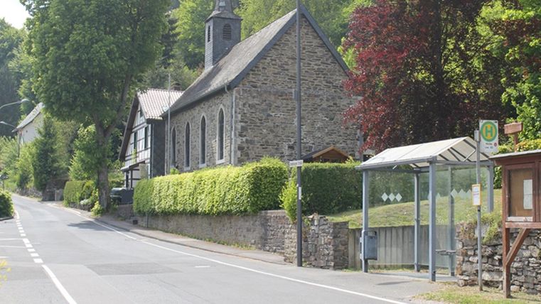 Een rustige straat met een stenen kerk en groene bomen. Aan de zijkant bevindt zich een bushalte.