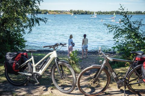 Deux vélos sont au bord d'un plan d'eau. À l'arrière-plan, deux personnes regardent l'eau et les bateaux.