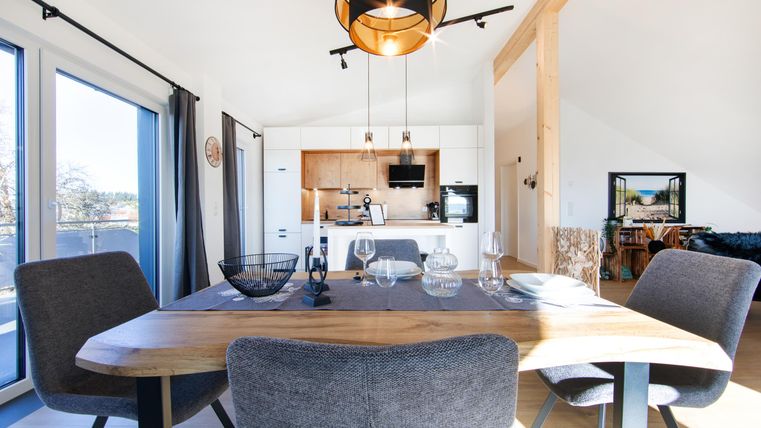 Een moderne eetkamer met een grote houten tafel en stijlvolle stoelen. Op de achtergrond is de open keuken in lichte kleuren,