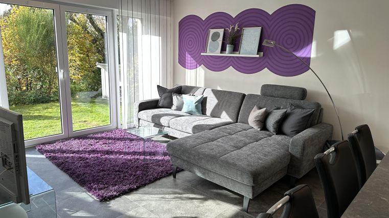 A modern living room with a comfortable gray sofa and a purple rug. Large windows provide plenty of natural light and a view of the greenery.