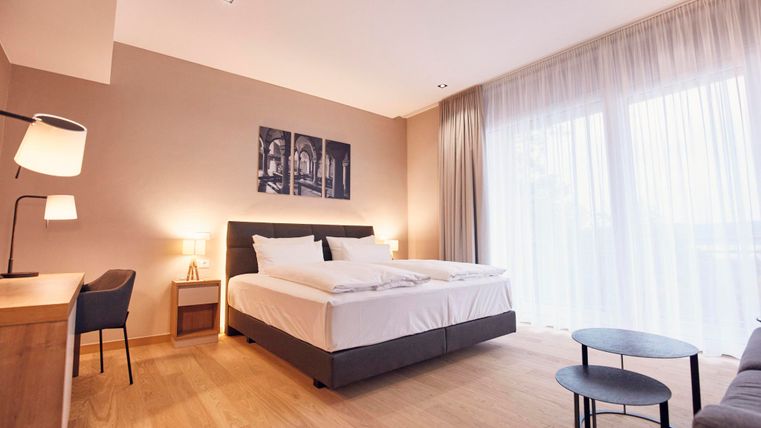 Een moderne hotelkamer met een groot tweepersoonsbed en een houten vloer. De kamer is helder en vriendelijk met grote ramen en stijlvolle meubels.