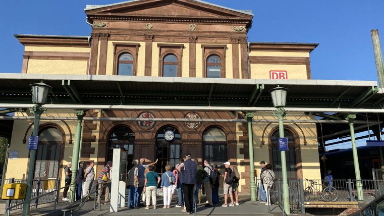 A historic train station with a distinctive facade. Numerous people are gathered in front of the building.