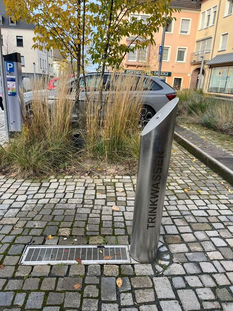 Ein Trinkwasserbrunnen aus Edelstahl steht auf einem gepflasterten Gehweg. Im Hintergrund sind Pflanzen und Gebäude zu sehen.