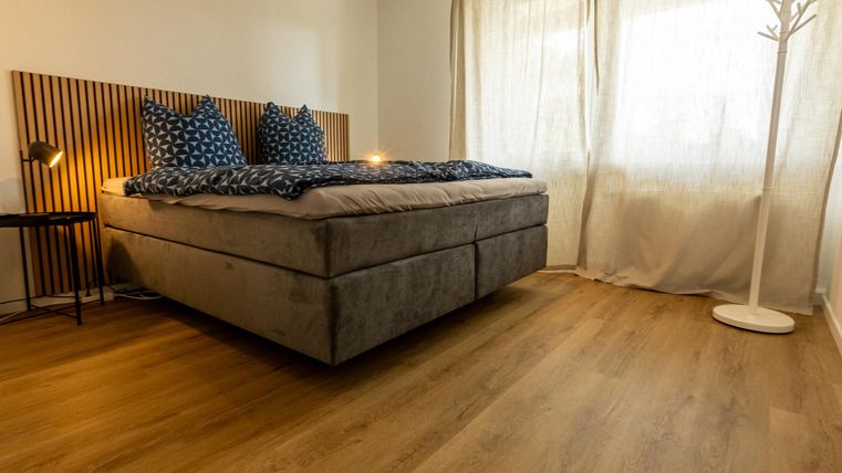 Een moderne slaapkamer met een groot bed en blauwe kussens. De ruimte heeft lichte muren, grote ramen en een warme houten vloer.