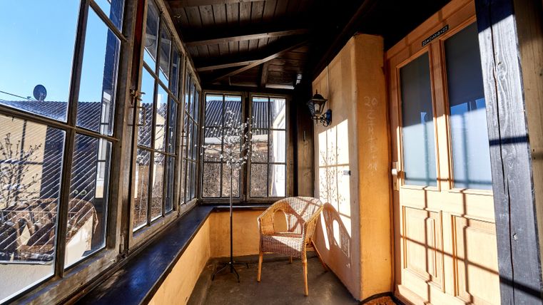 Ein heller Balkon mit großen Fenstern und einem Holztisch. Ein einzelner Stuhl steht vor der Tür und es scheint, als ob die Sonne scheint.