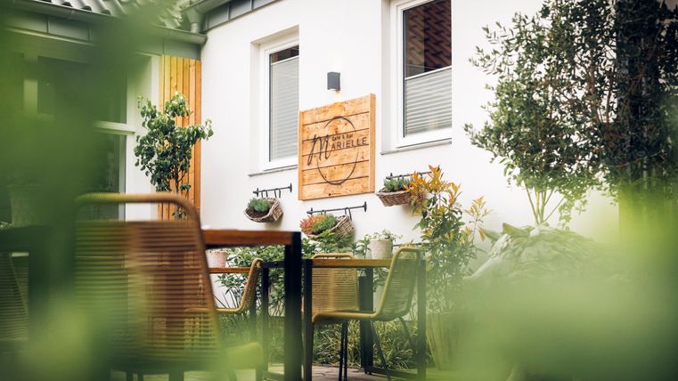 A cozy outdoor area with tables and chairs. In the background, there is a wooden sign with the name of the establishment and plants visible.