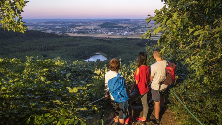 Drei Wanderer genießen den Blick auf eine malerische Landschaft mit Wäldern und einem See. Die Sonne geht langsam unter und taucht die Szenerie in warmes Licht.