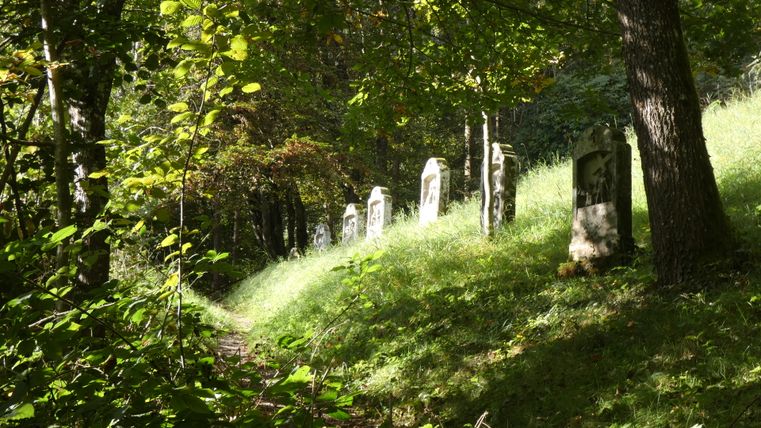 Ein schattiger Waldweg mit mehreren steinernen Kreuzwegstationen am Hang.