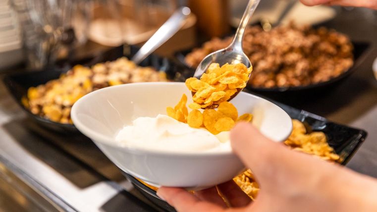 Een persoon vult cornflakes in een kom met yoghurt. Op de achtergrond zijn verschillende muesli en ingrediënten in kommetjes te zien.