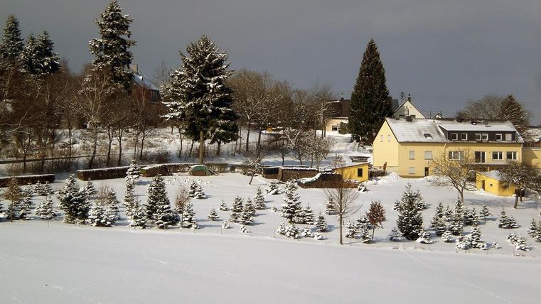 A snowy landscape with a yellow house and several small fir trees. The sky is gray and clouds are visible.