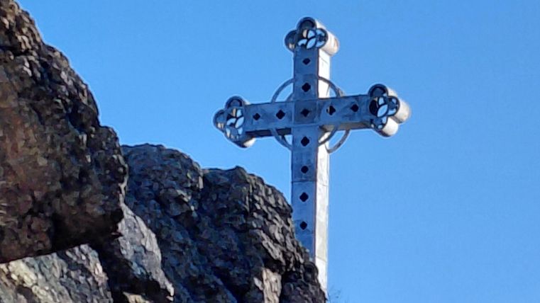 Une grande croix argentée se dresse sur un rocher sous un ciel bleu clair. L'environnement est vert et rocheux.