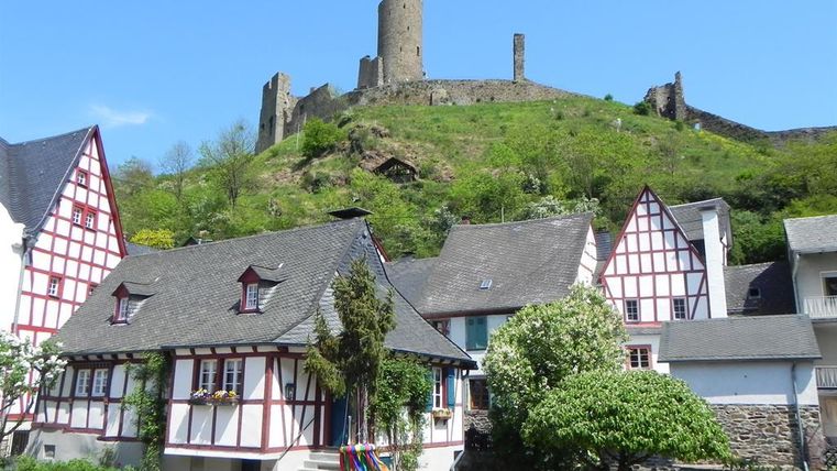 Eine malerische Landschaft mit traditionellen Fachwerkhäusern im Vordergrund. Im Hintergrund erhebt sich eine Burgruine auf einem grünen Hügel.