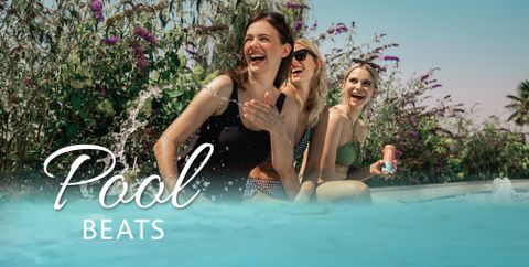Three cheerful women are sitting by the pool and laughing. Colorful flowers surround the relaxed atmosphere.