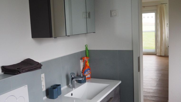 Ein modernes Badezimmer mit einem Waschbecken und einem großen Spiegelschrank. Der Blick führt durch die Tür in einen hellen Wohnraum.