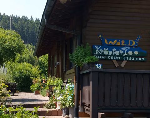 Ein gemütliches Holzhaus mit einem begrünten Vorgarten. Das Schild zeigt den Namen "Wild KräuterFee" und die Telefonnummer.