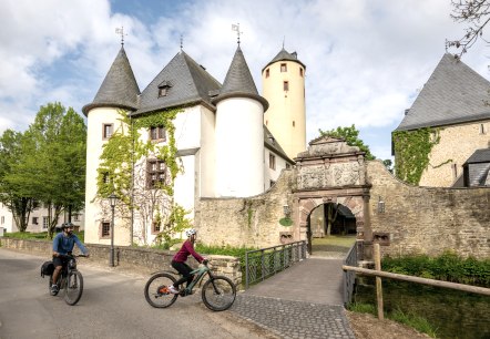 Zwei Radfahrer fahren an der historischen Burg Rittersdorf vorbei, die von grünen Bäumen und einem klaren Himmel umgeben ist., © Eifel-Tourismus GmbH/Dominik Ketz