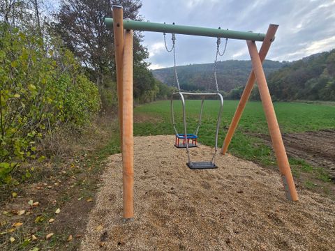 Eine Schaukel steht auf einem gepflasterten Sandboden in einem ruhigen Park. Im Hintergrund sind Bäume und sanfte Hügel zu sehen.