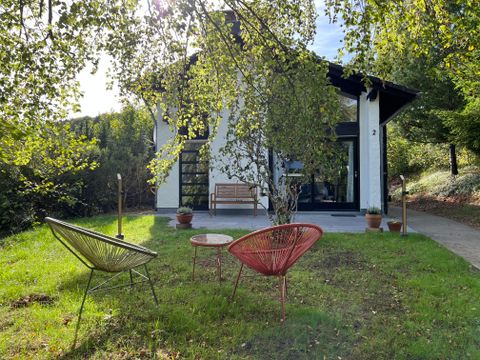 Ein modernes Haus umgeben von Bäumen und grünem Rasen. Vor dem Haus stehen zwei stylische Stühle und ein kleiner Tisch.