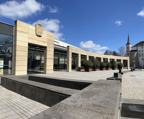 Ein klarer Platz vor einem modernen Gebäude mit einer schönen Pflanzenarrangement. Die Sonne scheint und der Himmel ist blau.