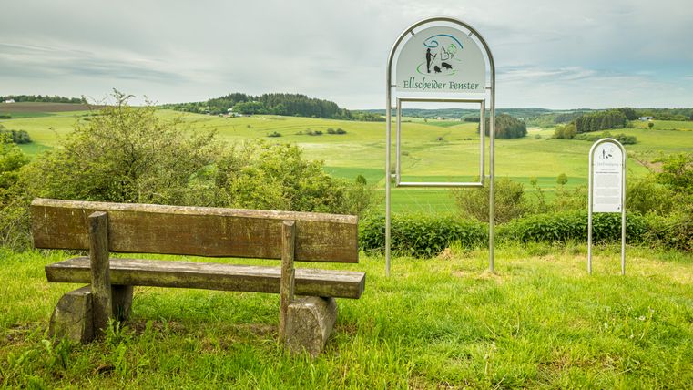 Wooden bench and sign with the inscription 'Ellscheider Fenster' in front of a green landscape.