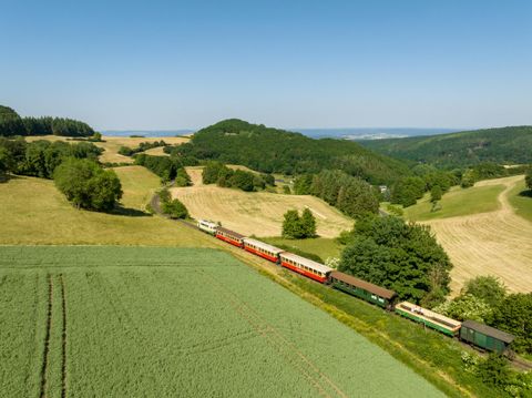 Een kleurrijke stoomlocomotief rijdt door een groene landschap. Op de achtergrond zijn zachte heuvels en een blauwe lucht te zien.