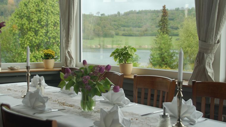 A beautifully set table with a floral arrangement and candles. Through the window, a green landscape with a body of water is visible.