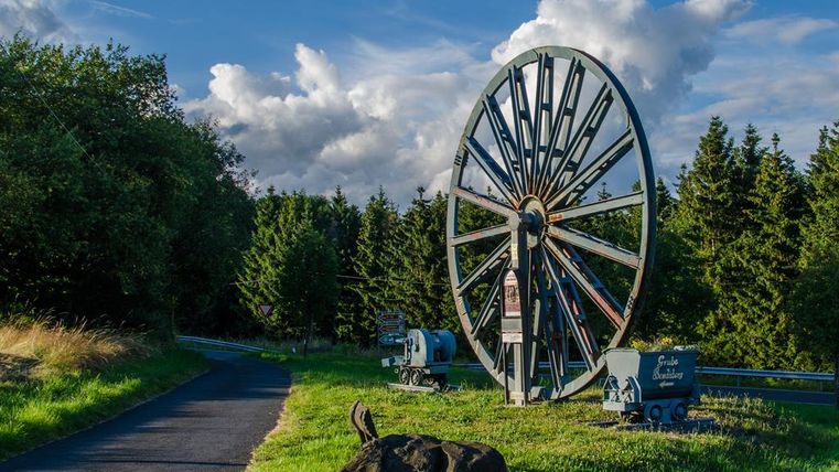 A large wheel stands on a meadow, surrounded by trees. The sky is blue with some clouds and a hiking path leads by.