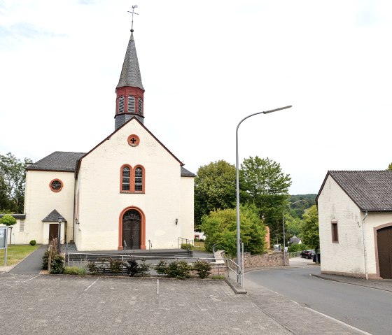 A church in Wißmannsdorf with a pointed tower, surrounded by trees. In front of it an empty parking lot and a road. Another building on the right., © TI Bitburger Land