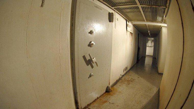 Een smalle gang met een massieve, gesloten deur aan het einde. De vloer lijkt vuil en de muren zijn eenvoudig en wit geschilderd.