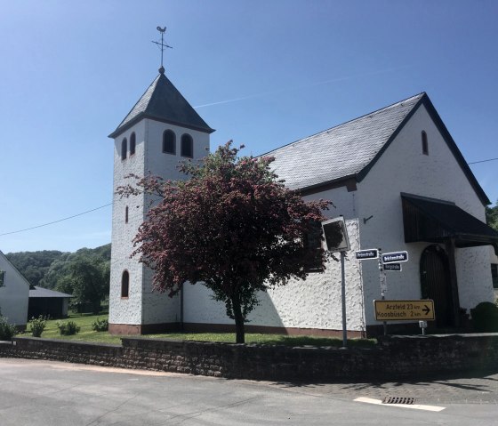 White church with tower and red tree in the foreground, blue sky. Road signs point to Arzfeld and Koosbüsch., © Fellmann
